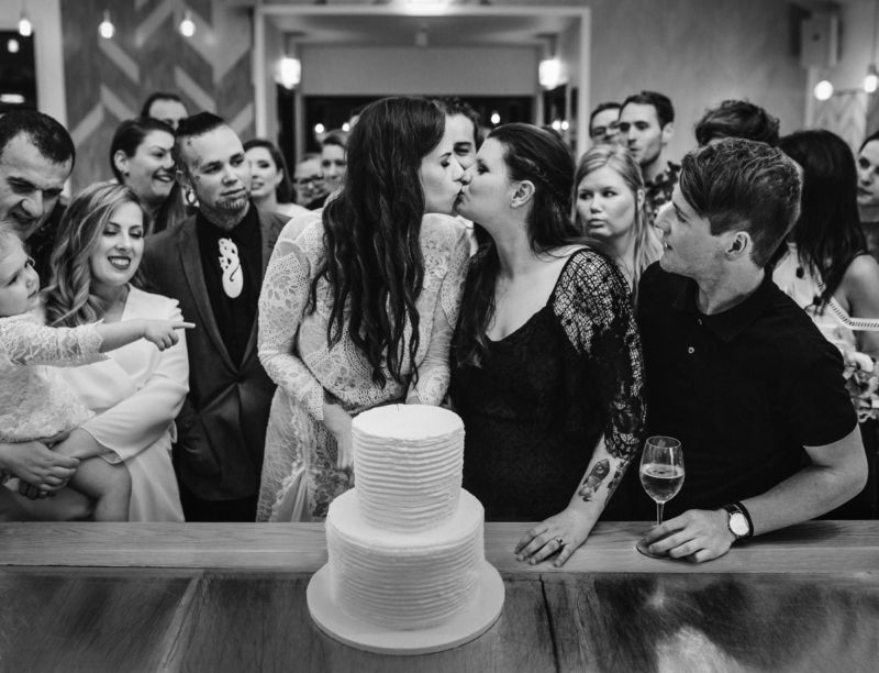 brides kissing and cutting cake surrounded by loved ones