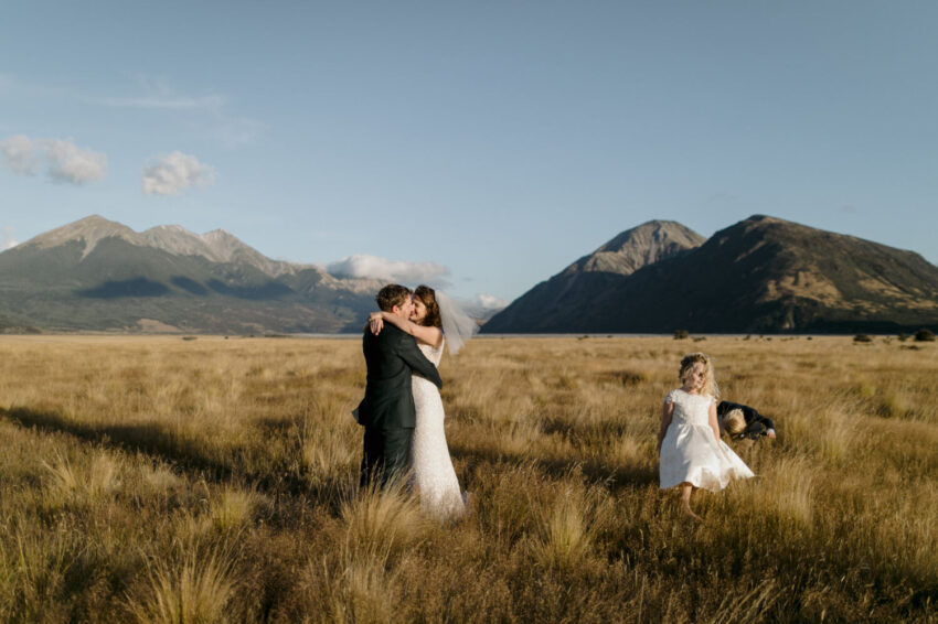 Bride and Groom kissing in field with children running up to them