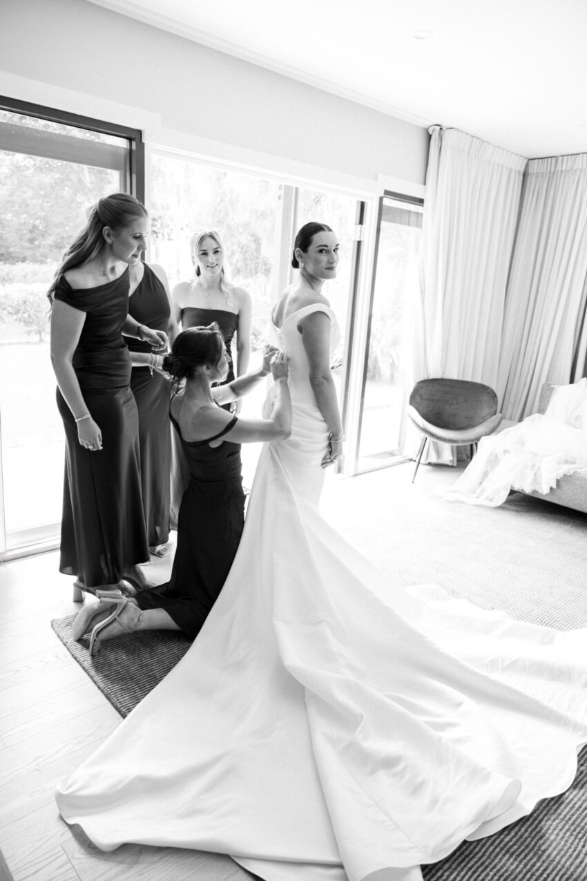 Bride looking right at camera as bridesmaids help her get dressed