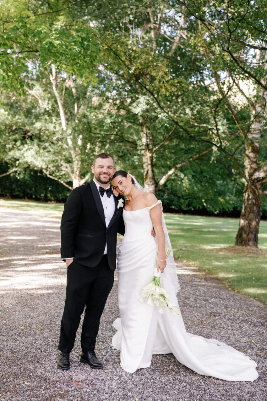Bride and Groom standing in a driveway of trees smiling for a portrait