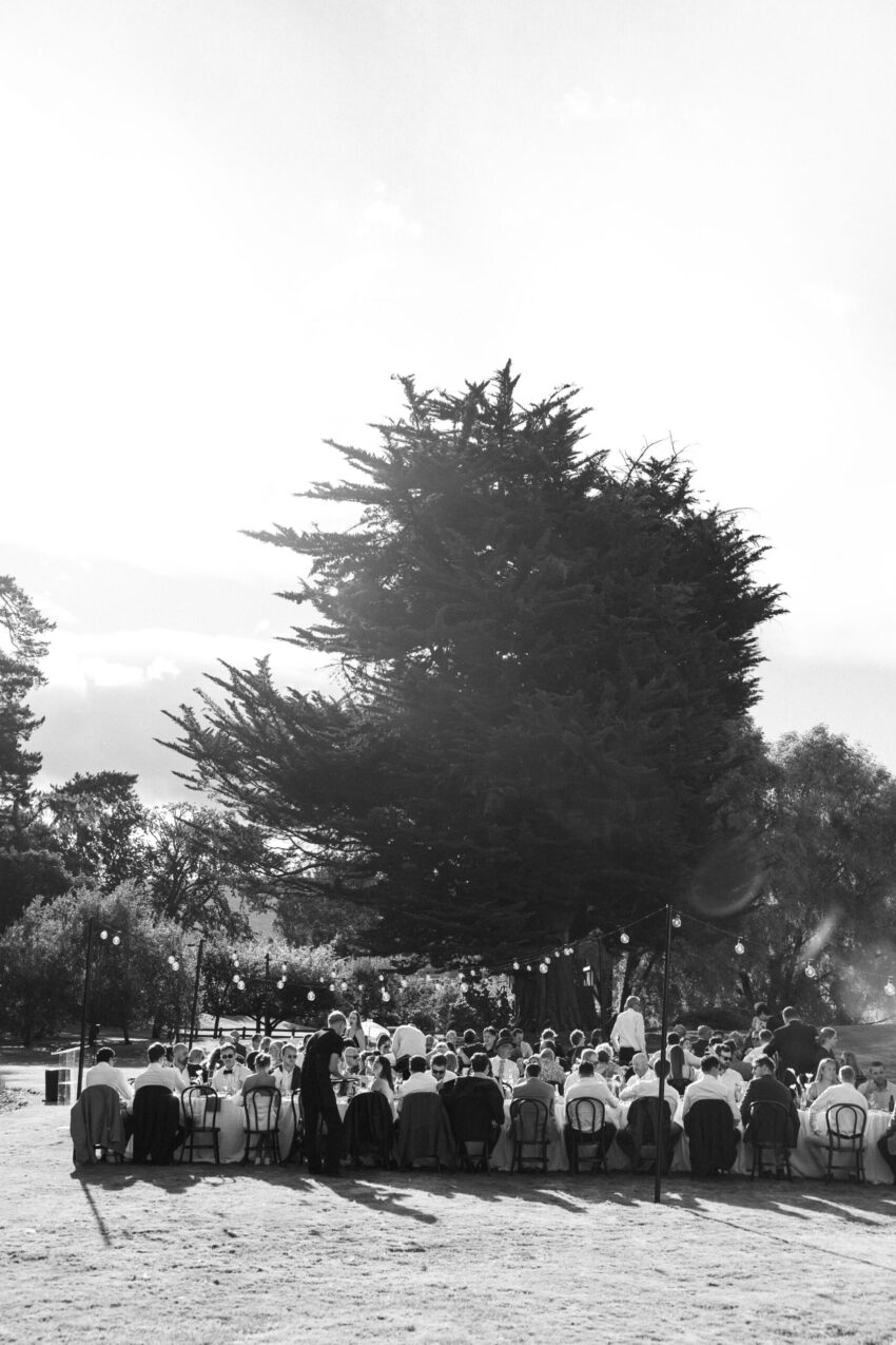 wide B&W of the outdoor reception with guests sitting at long tables with bulb lighting hanging over the setting
