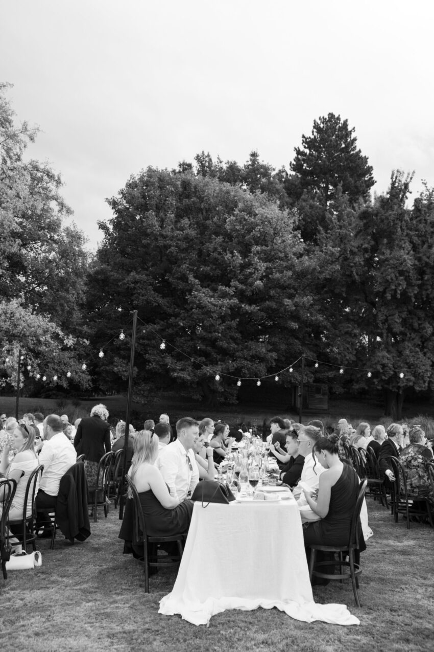 B&W of the outdoor reception with guests sitting at long tables with bulb lighting hanging over the setting
