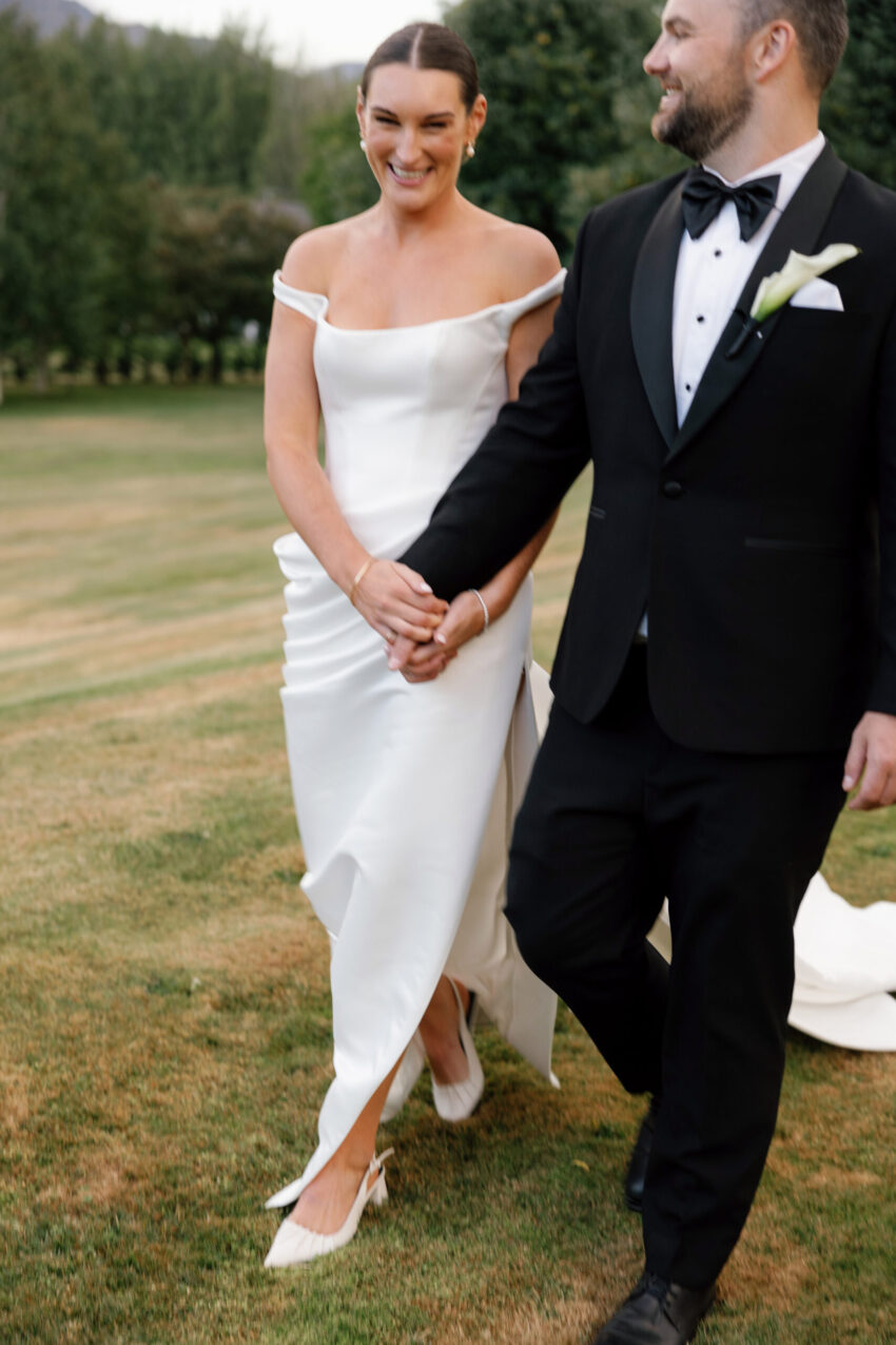 Colour photo of Bride smiling at the camera and she holds the grooms hand and walks along