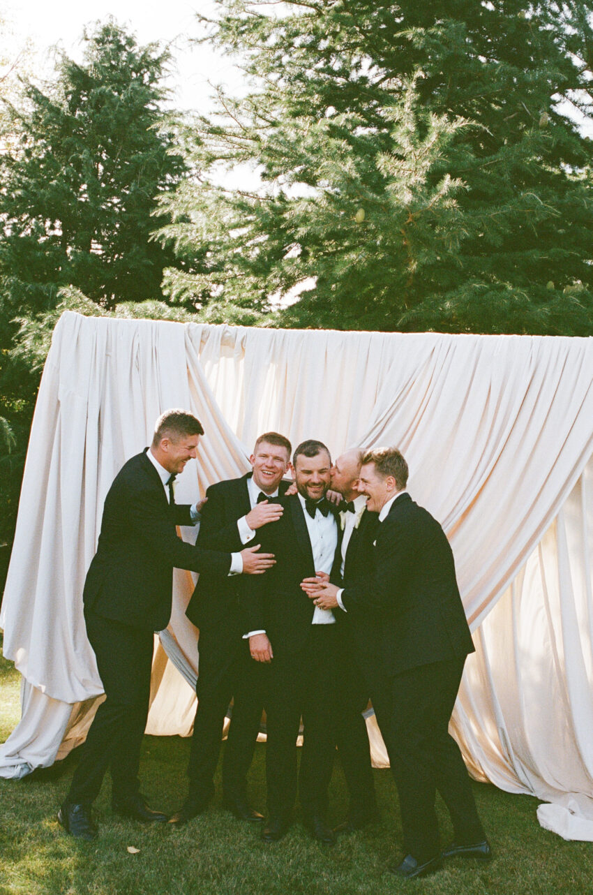 Groom being hugged and kissed by the groomsmen in front of a draped backdrop