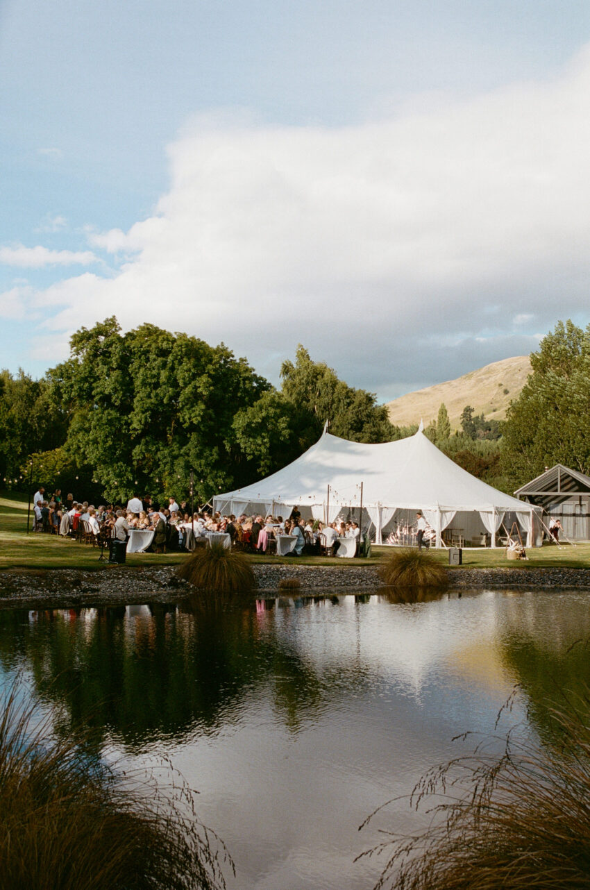 The reception scene across a pond giving a slight reflection of the tent and the blue sky